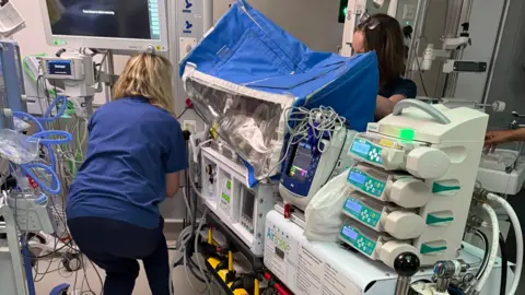 Jade A hospital scene showing a specialised transport incubator on a wheeled stretcher, equipped with multiple infusion pumps and monitoring devices. A healthcare professional is adjusting equipment nearby with another person partially visible behind the incubator.