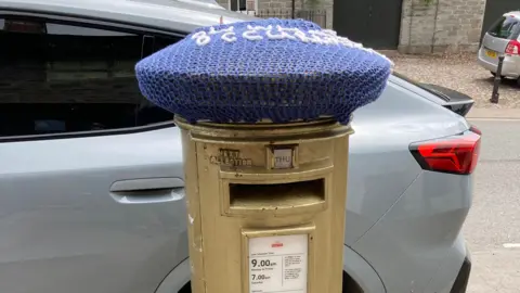 The gold postbox has a car parked behind it and the remains of the blue postbox topper on top.