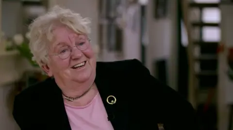 BBC Nell McCafferty in 2016, smiling and looking out of frame. She wears glasses, a black blazer, a pink top and a gold brooch.