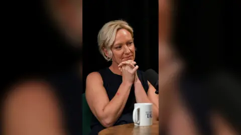 Ruth Dodsworth clasps her hands together by her chin. She is wearing a black sleeveless top and looking to her left.