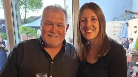 A man on the left with grey hair and a dark shirt is holding a glass of drink in his hand. A woman with long brown hair and wearing a dark top is next to him.