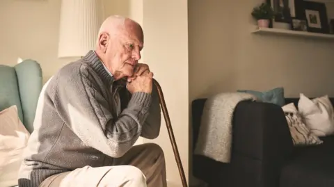 Getty Images An elderly man in a brown zipped cardigan sits on a green chair in a care home, leaning against his stick. There is a lamp and another sofa with cushions in the background as well as a shelf containing plants and pictures.
