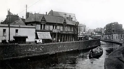 Getty Images A black and white photo of a canal, with walls and buildings either side and a single canal boat on it.
