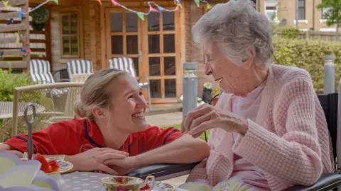 A women in a red top is kneeling next to an older woman sat down. There is afternoon tea in front of them and bunting in the background. 