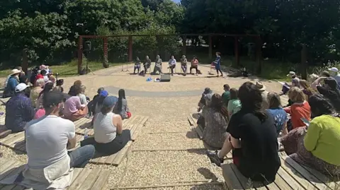 Birmingham Settlement An amphitheatre with a performance being watched by audience who are sitting looking at the performers with their backs to the camera.