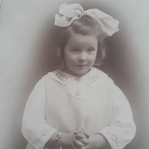 Bob Lane An old black and white picture of a girl with her hair tied up in a large white bow and wearing a white gown.