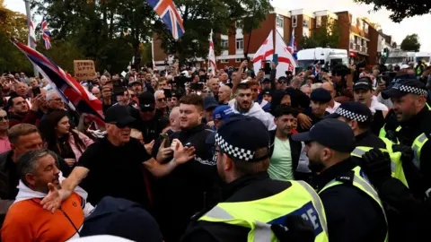 Reuters A line of police officers face up to a large crowd of people who appear to be protesting. Some are waving St George and Union Jack flags in a town centre setting.