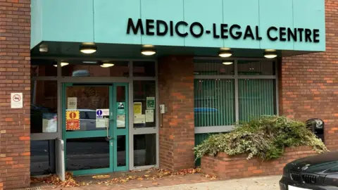 Oli Constable/BBC A blue-sign saying Medico-Legal Centre above a blue-framed door in a red-brick building.