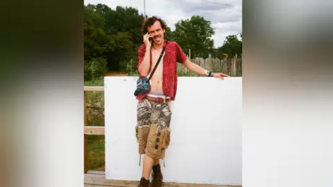 Eji Bazunu Woody Cook stands in front of a white wall. He's wearing an open red shirt and camouflage shorts
