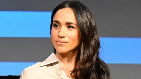Getty Images Meghan, who has long wavy brown hair, has a neutral expression as she looks on during a talk on stage