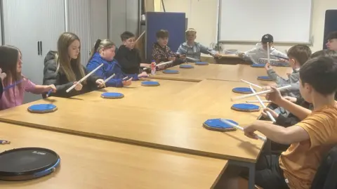 Around a dozen children are sitting around a number of tables that have been pushed together. In front of them are drum pads and each child has two drum sticks which they are using to strike the plates 