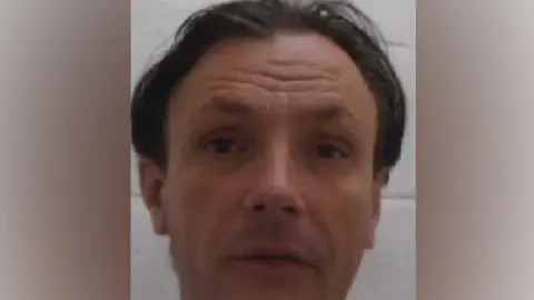 A Marcus Yeldham mugshot that is fairly blurry. He has dark, ear-length hair parted in the middle and has both his eyebrows raised