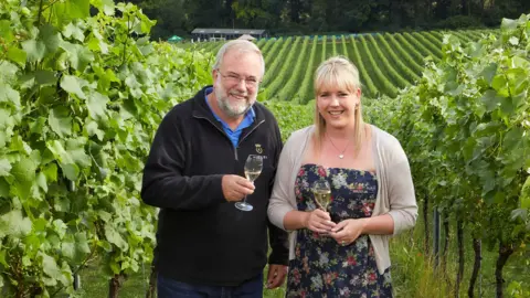 Nick Wenman and Lucy Letley stood in between rows of vines. The rest of the field can be seen down the hill behind them. They are holding full sparkilng wine flutes and smiling.