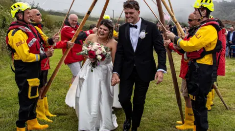 The newlyweds are smiling as they walk through a guard of honour of a lifeboat crew dressed in their kit, including their distinctive yellow wellies