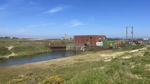 A general view of the Benacre sluice. A large green field can be seen with a river running through the middle of it. A pylon can be seen in the distance as well as two wind turbines and a small red brick building.