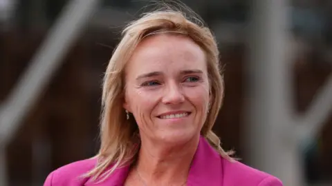 Sandie Peggie smiles and looks off to the side. She has shoulder length blonde hair and wears a pink blazer.