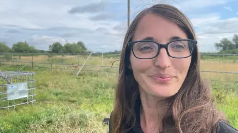 A woman with brown hair and glasses, stood in a grassy field.