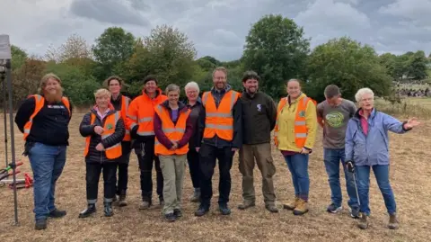 11 people, many wearing high visibility jackets, are standing in a line looking at the camera They are standing on dry ground in front of a row of trees