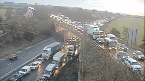 A traffic camera shows the A14 and A11 running over the top with long queues of vehicles. The majority of the vehicles have their headlights on as they wait in the queue.