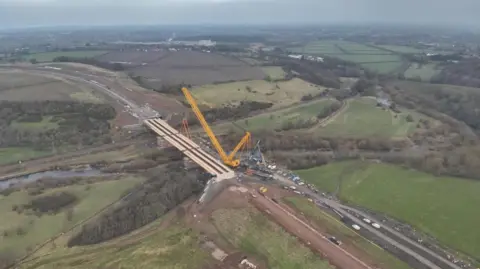 Cumberland Council Construction work being carried out on the Caldew Crossing. Cranes are lifting material onto the unfinished bridge. The bridge stands over the River Caldew. It is surrounded by trees and woodland.