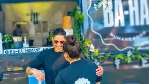 Two people stand in front of a food van decorated in florals which says 'BA-HA' on it.