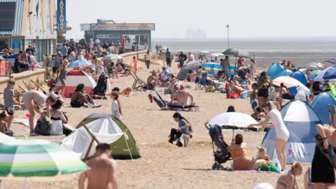 PA Media People on a sandy beach. There are small tents and beach umbrellas across the sand. In the background the sea is visible to the right and to the left is an arcade.