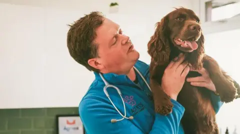 ACE Vets A still image of James Buick holding a brown cocker spaniel dog in an examination room. He has short, dark hair and is wearing a blue zip up fleece. He has a stethoscope around his neck.