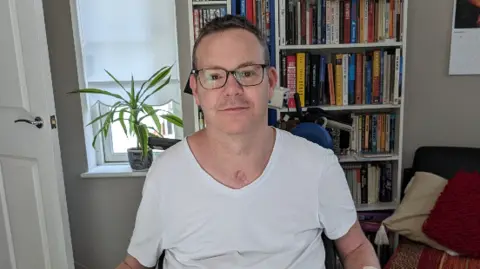 A man with cropped hair and glasses in a white T=shirt. 