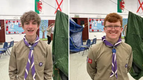 BBC/RICHARD EDWARDS These pictures show two explorer scouts, Daniel and Noah, at a weekly unit meeting in Copmanthorpe, York. Daniel on the left has curly brown hair and is waering a khaki-coloured scout uniform shirt and a purple and white neckerchief. Noah on the right has short red hair is wearing the same uniform as Daniel, and is also wearing glasses. They are are both smiling and are standing in the hall used by the local scouts for their regular get-togethers.