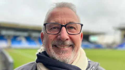 Emma Baugh/BBC Michael Ferguson is smiling at the camera while stood inside the Weston Homes Stadium. The pitch behind him is empty, as are the blue seats in the stands. Michael has grey facial hair and grey hair as well as black square framed glasses.  He has a blue and white scarf wrapped around his neck. 