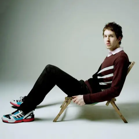 Getty Images Calvin Harris with very dark hair. He is sitting in a low deckchair, wearing black jeans, trainers and a brown jumper. 