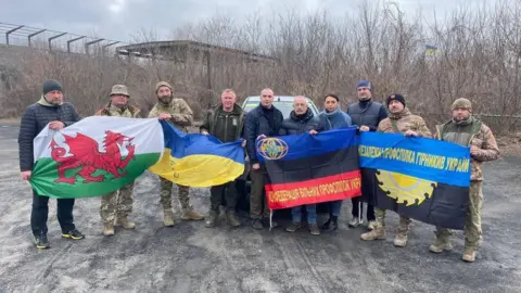 KVPU, the Independent Union of Ukrainian Miners A group of men stood in a row behind four different flags, including a wales flag and a Ukraine flag. The other flags are black, blue and red, and black and blue, and contain Cyrillic writing. 