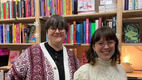 There are two women stood in front of book shelves in a shop. The woman on the left has short brown hair and glasses, she is smiling and wearing a black top and a red and white cardigan. The woman on the right is also smiling, she has shoulder length brown hair and glasses and is wearing a cream jumper