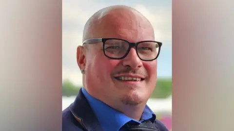 Handout A man smiles at the camera. He is bald and has a goatee beard. He is wearing a blue shirt, cravat and denim jacket and is wearing black framed glasses.