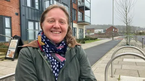 A woman is standing outside in a green jacket an blue and white scarf. She looks happy.
