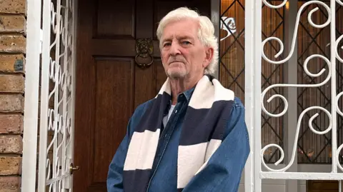 Anthony Burton, a man with white hair and a dark denim shirt, stands outside his home looking disgruntled.