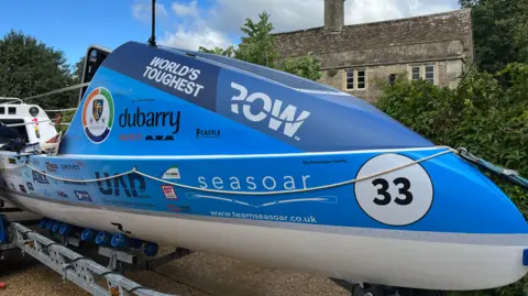 A rowing boat is pictured from the front. It has a small, indoor space with a sloped roof that looks barely large enough for two people to lie down side by side.