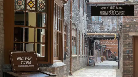 A walkway through a 19th Century pottery factory. A window on the left has a brown sign underneath that has "Middleport Studios" written on it. Light bulbs are hung up across and above the walkway.