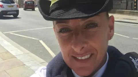 Handout A middle aged woman wearing a traffic warden's uniform, including a black hat and white shirt, smiles as she takes a selfie on a pavement by a busy main road.