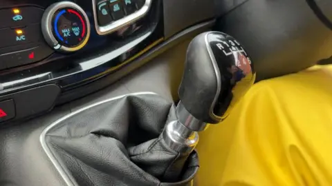 An up-close view of a manual gear stick. In front of the stick are climate controls for the vehicle. The person driving is wearing yellow clothing.
