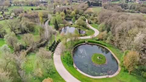 Fine and Country A pond in the foreground is surrounded by trees with a snaking path going around the outside.