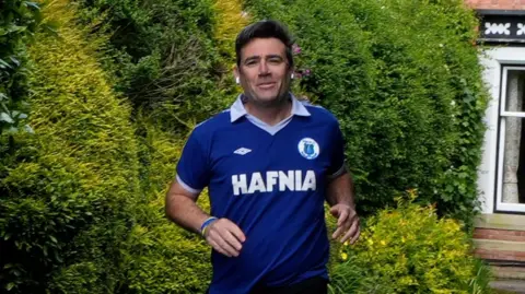 PA Media A man a blue sports shirt running towards the camera with green bushes behind him and a window from a house on the right.