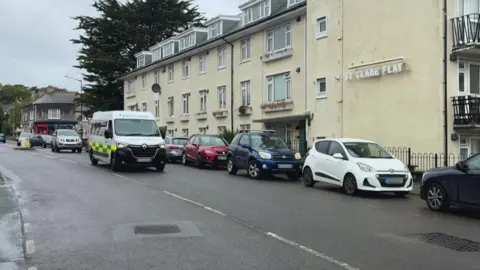 A white ambulance drives down a road in Penzance which has several cars parked along the side of it. A block of flats called 'St Clare Flat' is on the road.