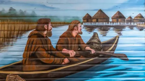 Historic England Archive A mural showing an artist's impression of two men in a log boat on water and heading to a floating settlement of thatched huts on the horizon. The men have full beards, are wearing brown clothing and sitting one behind the other. The lead man has both hands on a paddle, dipping into the blue water. They are looking towards the settlement. Over their shoulders is an extensive bed of reeds. 