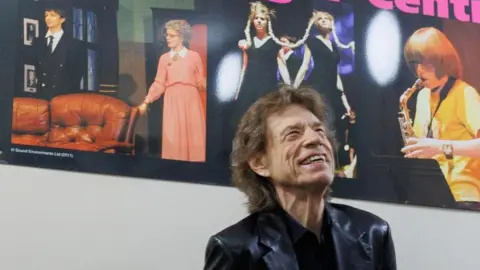 Pete Bresser An elderly man in a leather blazer. He has long hair and is smiling. Behind him is a poster of musicians. 