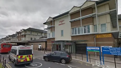 External view of the Royal Oldham Hospital showing the A&E department and an ambulance, bus and car driving past the entrance 