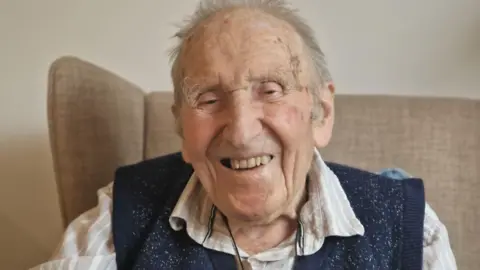 Sanctuary Care A man wearing a blue top and a grey and white shirt is smiling at the camera. He is sitting in a brown chair in front of a plain wall.