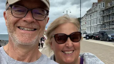 Sean Cleghorn Sean Cleghorn takes a selfie with his wife on a seafront promenade. He has a short brown and white beard and wears a cap, a grey T-shirt and glasses. Allison has straight, blonde hair, sunglasses and a grey top.