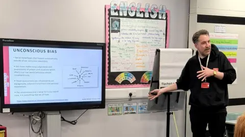 Show Racism the Red Card James Kingett, with brown curly hair and a beard, wears a black hoody, black trousers and a lanyard with his sleeves rolled up as he gestures toward a white board displaying a slide about 'unconscious bias' in a classroom
