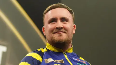 PA Media World Darts Champion Luke Littler with short brown hair wearing his purple and yellow darts shirt. He is standing near the oche with the stage in the background. He is smiling. 